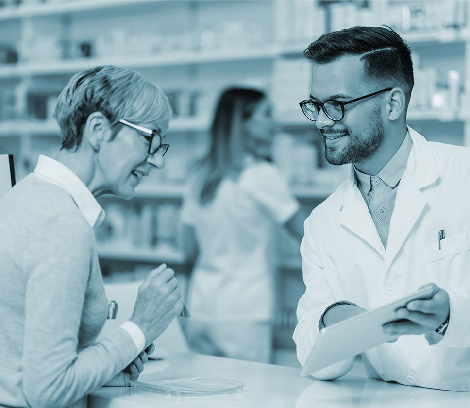 A photo of a bearded pharmacist consulting with a female patient at the pharmacy counter